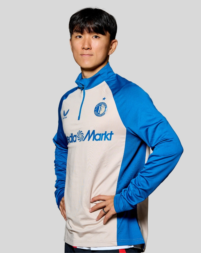 Male model wearing blue and beige Feyenoord stadium midlayer top with crest and sponsor on chest.