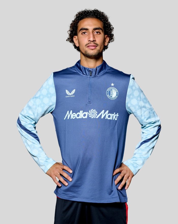 Male model wearing Feyenoord Uit Matchday Midlayer Top 2025-2026, front view showing dark blue body, light blue sleeves and sponsor logos.