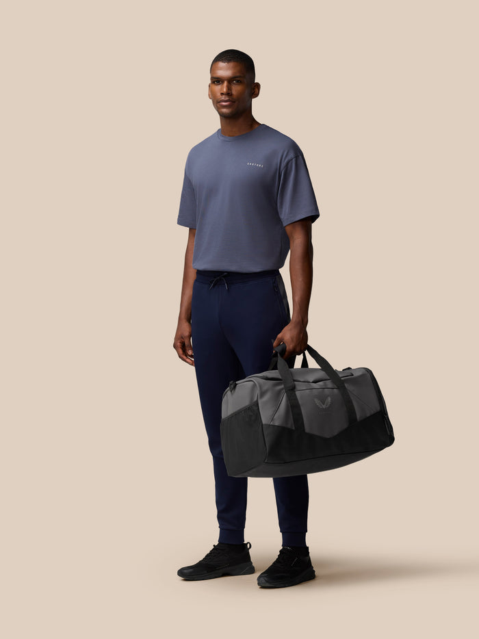 Male model with gray and black Castore Core Holdall duffel bag in hand