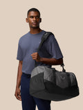 Unisex Core Holdall in gunmetal and black carried over the shoulder by a model against a beige backdrop.