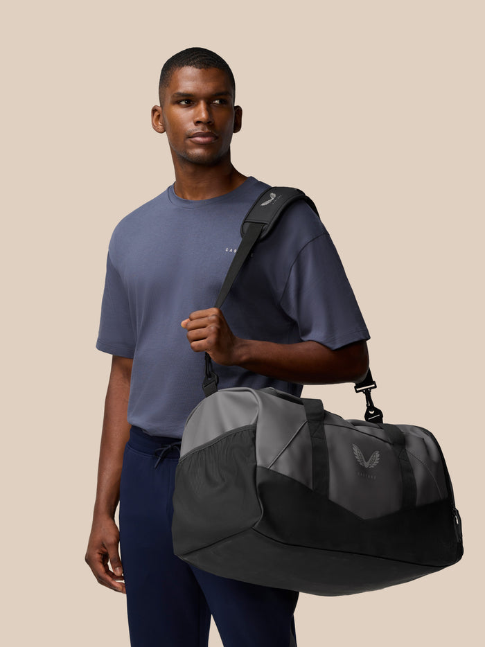 Unisex Core Holdall in gunmetal and black carried over the shoulder by a model against a beige backdrop.