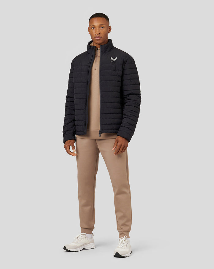 Full body shot of a man in a black Castore puffer jacket over a beige hoodie and tan joggers with white sneakers