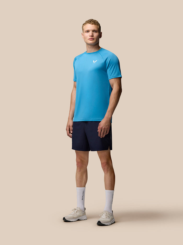 Full body shot of a male model in blue Adapt T Shirt with a white Castore logo on the left chest, navy shorts and white sneakers.