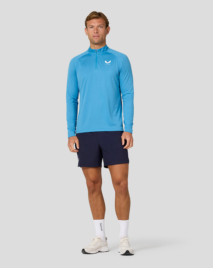 Full-length shot of a man wearing blue long sleeve top with quarter zip and Castore logo, navy shorts