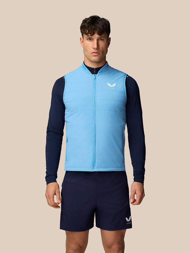 Apex Training Gilet in lichtblauw gedragen door mannelijke model, mouwloos gewatteerd vest met ritssluiting en Castore-logo op de borst