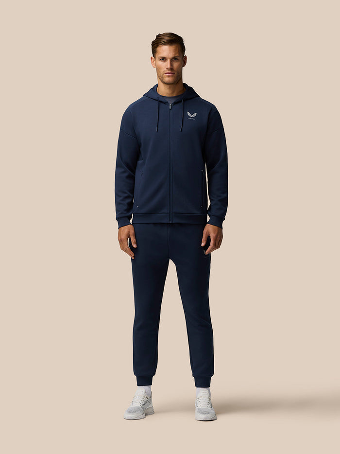 Full body shot of a man in navy Travel Lifestyle Joggers and hoodie set on a beige background