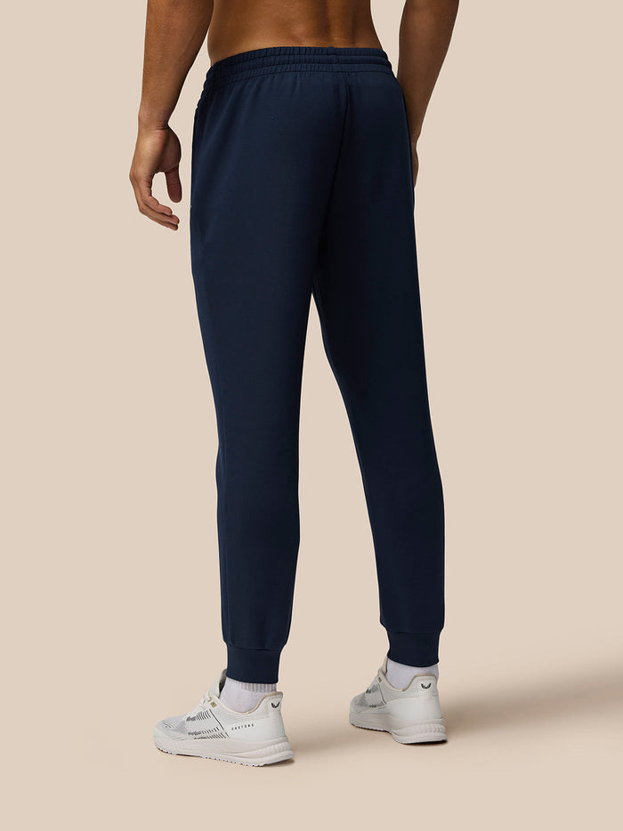 Front view of navy Travel Lifestyle Joggers with logo on the thigh and ribbed cuffs paired with white sneakers.