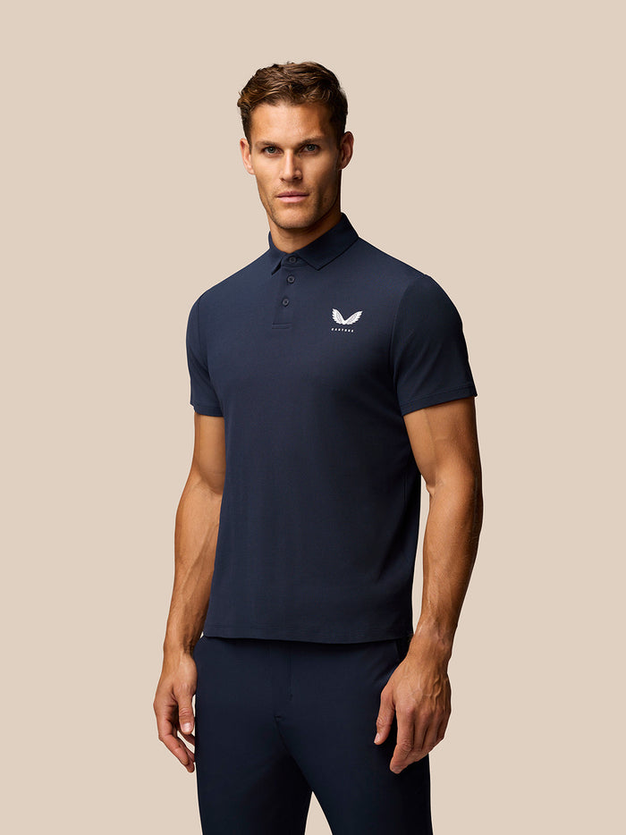 Male model wearing dark blue Golf Club Essential Polo with Castore logo on chest, standing against a beige background