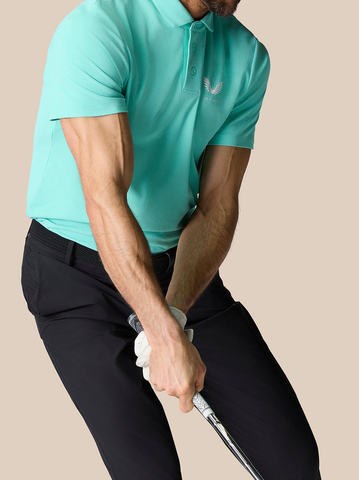 Side view of a man wearing a light aqua Golf Club Essential Polo while swinging a golf club with the logo visible on chest