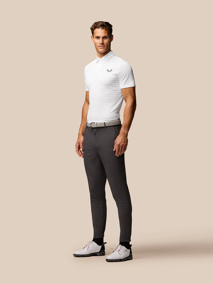 Full body of a male model in a white Golf Pinnacle Pique Gestreepte Polo with grey trousers and white golf shoes.