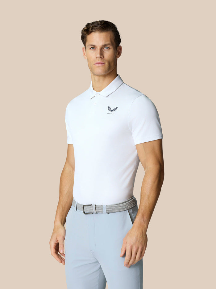 Male model wearing white Golf Pinnacle Polo with light blue pants and a gray belt against a beige backdrop.