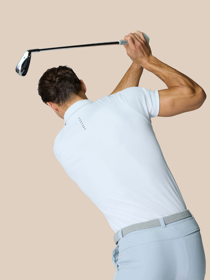 Male golfer in a white polo and light trousers performing a backswing with the club raised over his head