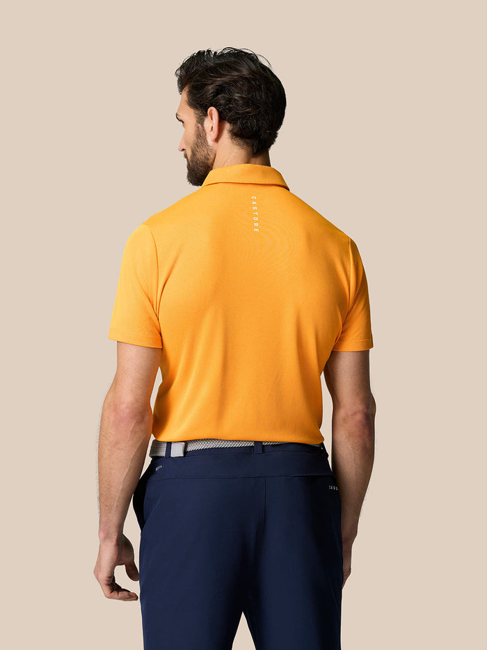 Back view of a man wearing a Mango orange knit polo with short sleeves and a Castore logo on the upper back against a beige background.