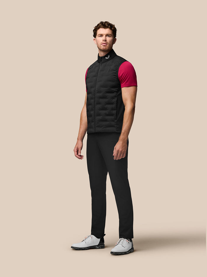 Volledig beeld van een man in een zwarte Pinnacle Gilet over een roze t-shirt, staand met gesloten ritssluiting en opstaande kraag.