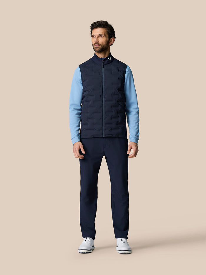 Full length view of a man wearing a navy Golf Pinnacle Gilet over a blue long sleeve top with a stand up collar