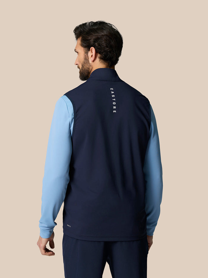 Back view of a man wearing a navy Pinnacle Lightweight Hybrid Gilet with light blue sleeves and vertical CASTORE branding along the spine