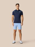Full-body view of a man in navy Pinnacle Golf Polo and light blue shorts standing against a beige backdrop