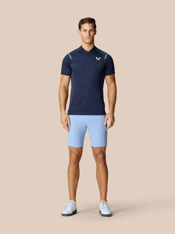 Full-body view of a man in navy Pinnacle Golf Polo and light blue shorts standing against a beige backdrop