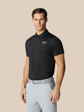 Model draagt zwarte grafische polo met golfbalprint en Castore-logo op borst, gecombineerd met lichtgrijze broek.