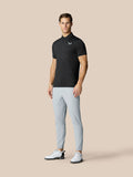 Volledig beeld van een man in een zwarte grafische golfpolo met puntige kraag, lichtgrijze broek en witte golfschoenen.