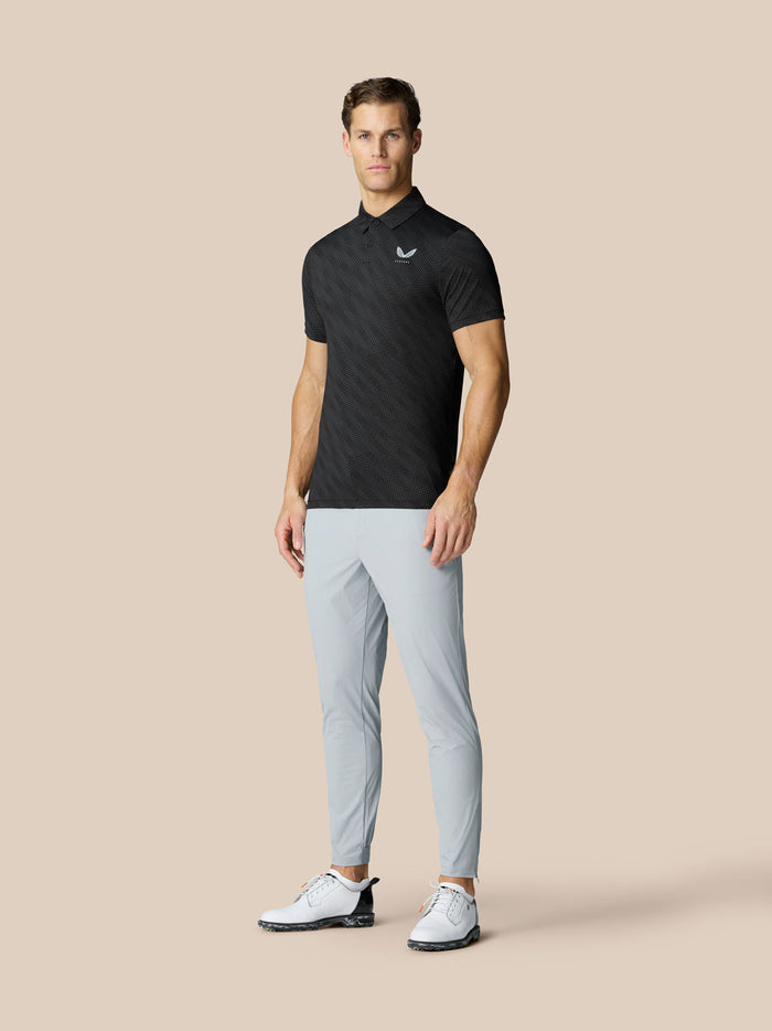 Volledig beeld van een man in een zwarte grafische golfpolo met puntige kraag, lichtgrijze broek en witte golfschoenen.
