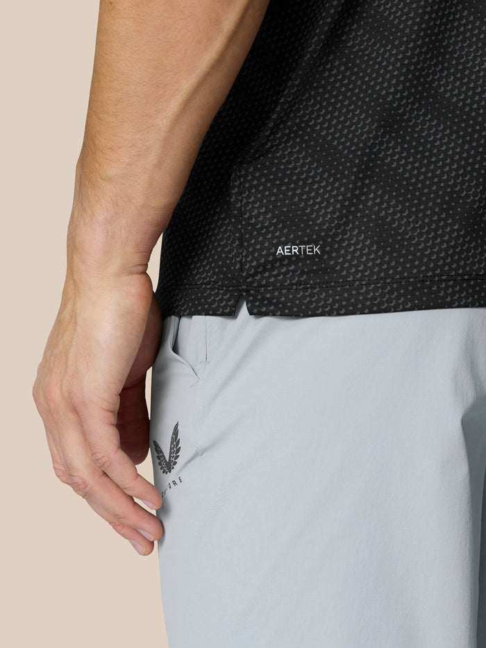 Close-up van de zijkant van een zwarte grafische golfpolo met AERTEK label, gedragen met lichtgrijze shorts.