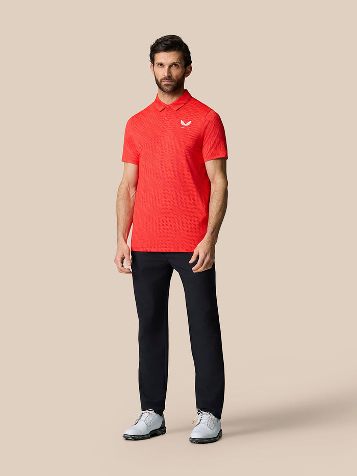 Male model wearing a red Golf Club Grafische Polo with a white logo on the chest, black pants, and white shoes against a beige backdrop.