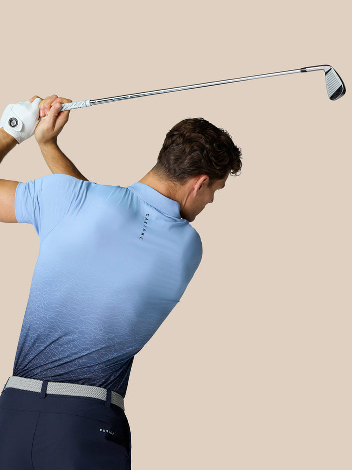 Golfer in een hemelsblauwe Club Golf polo met ombre print zwaait met een golfclub tegen een beige achtergrond.