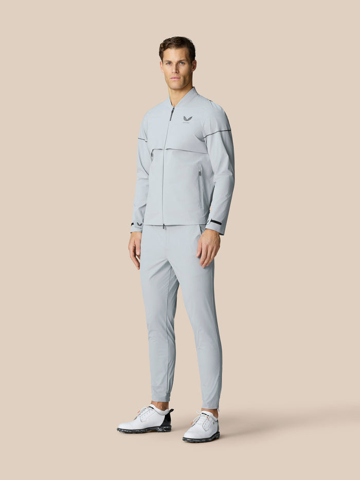 Full-body shot of a man wearing a matching light gray Golf Pinnacle woven jacket and pants with a chest logo and white golf shoes.