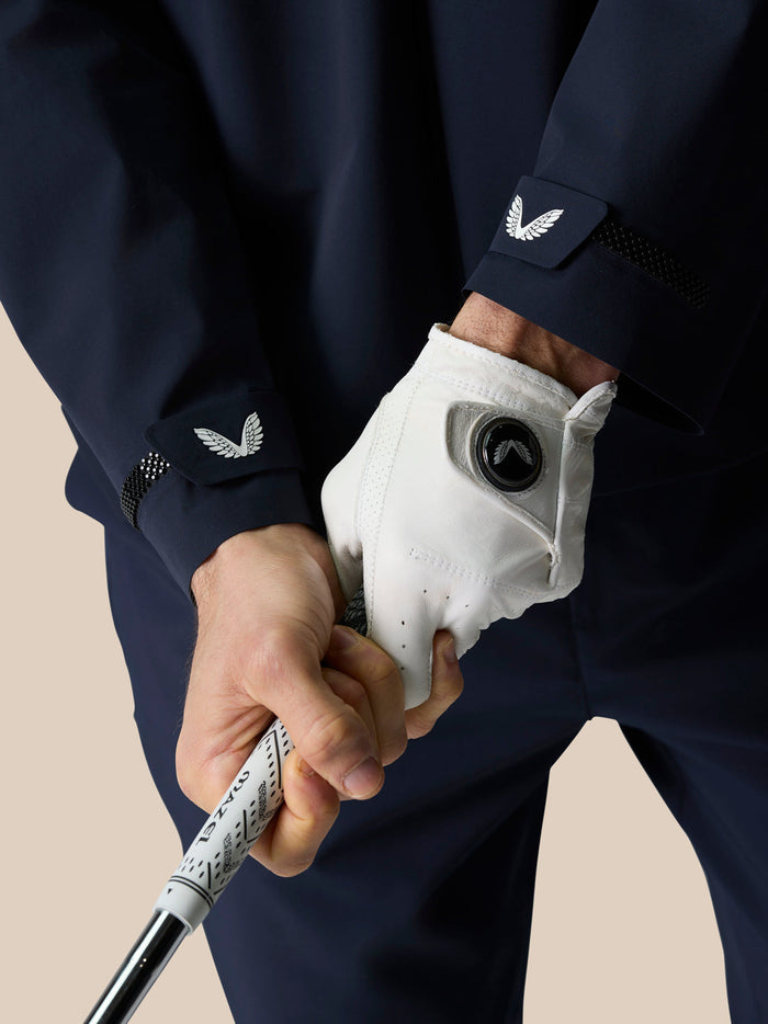 Close up of a golfer gripping a golf club in a navy Pinnacle jacket with a white glove and a wing logo on the cuff