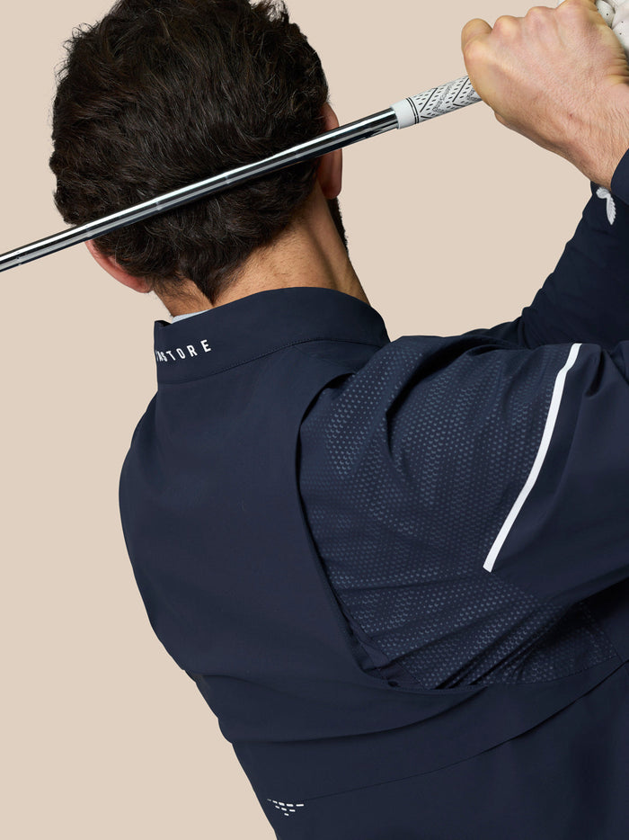 Rear view of model in navy Golf Pinnacle woven jacket with perforated sleeve panels and CASTORE collar branding while holding a golf club