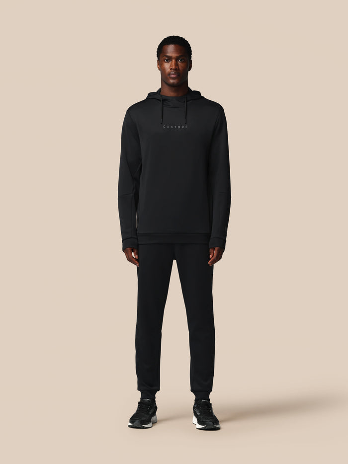 Male model wearing black Castore Flex long sleeve hoodie with hood against a beige background