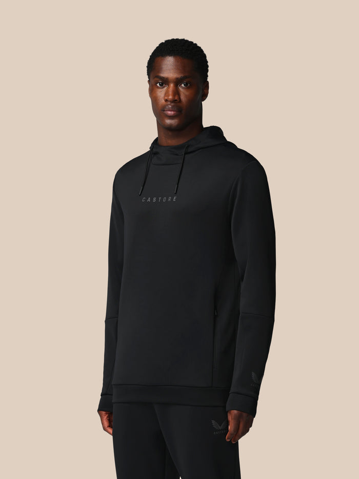 Full length view of a male model in a black Castore Flex hoodie with hood and chest logo against a beige background