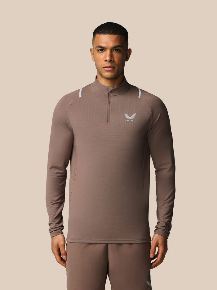 Male model wearing taupe Castore Apex quarter zip long sleeve top with raglan sleeves reflective shoulder accents and chest logo