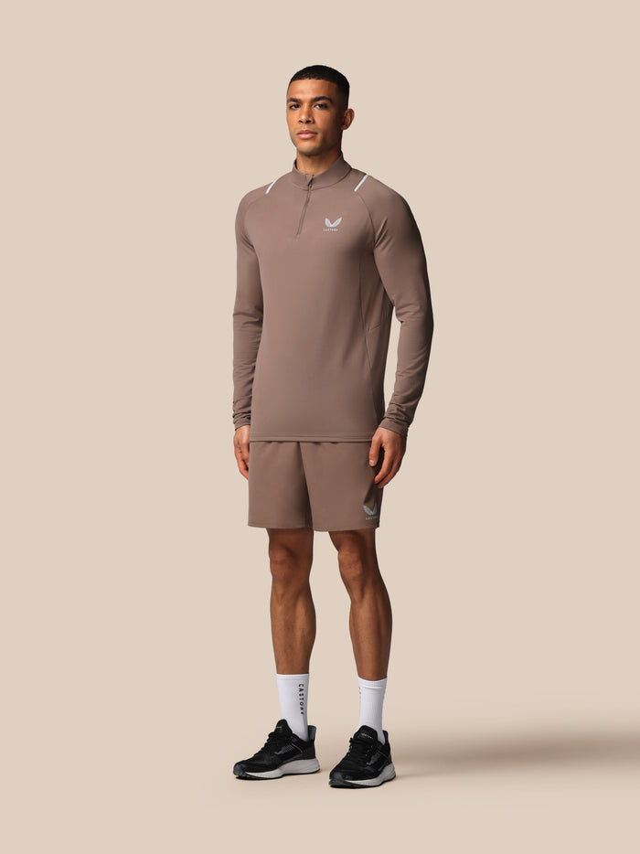 Front right view of male model in taupe apex training top with zip neck and matching shorts with Castore logo on chest and leg