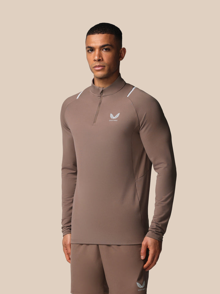 Male model wearing taupe Castore Apex training top with quarter zip and reflective shoulder accents